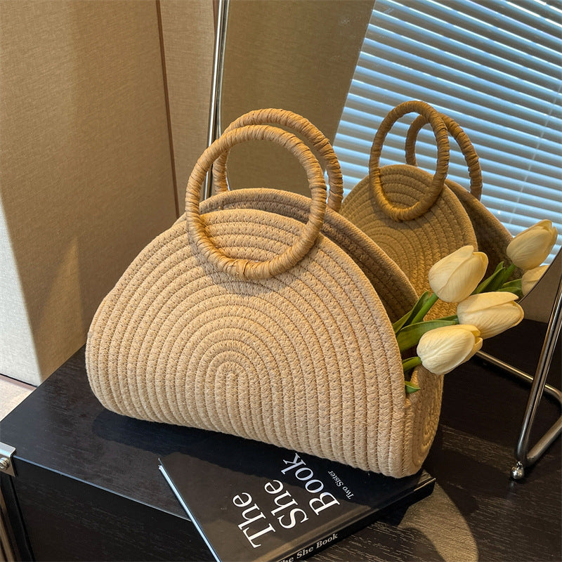 A woven handbag in a beige color with a shell shape and two handles, placed on a surface next to a bouquet of white flowers.