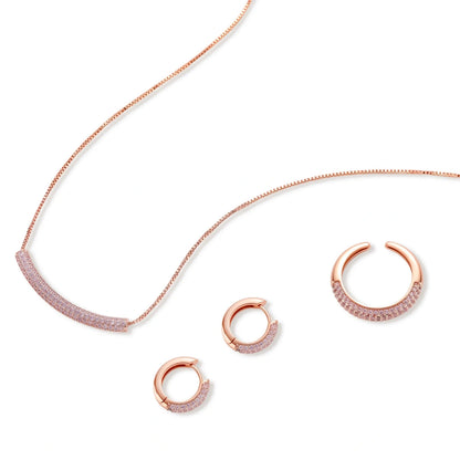 Rose gold jewelry set with pavé cubic zirconia stones including curved necklace, hoop earrings, and adjustable ring on cream background.