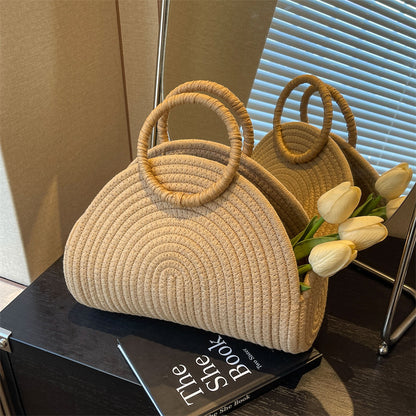 A woven handbag in a beige color with a shell shape and two handles, placed on a surface next to a bouquet of white flowers.