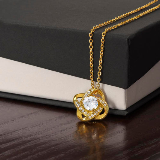 Gold love knot necklace with a sparkling cubic zirconia center, elegantly displayed against a polished wooden box background.