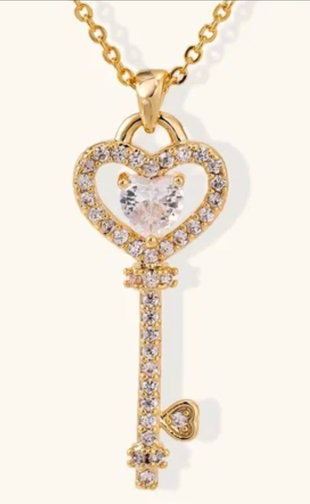 A gold key-shaped pendant necklace adorned with clear crystals, featuring a heart-shaped crystal centerpiece and a small dangling heart charm, shown on a light cream background.