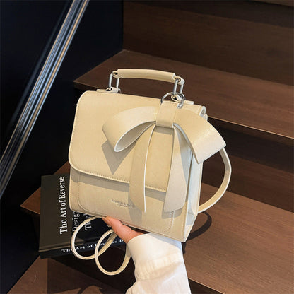 A creamy-white backpack with a bow design on the front, visible straps, and a zipper closure, held in a person's hand.