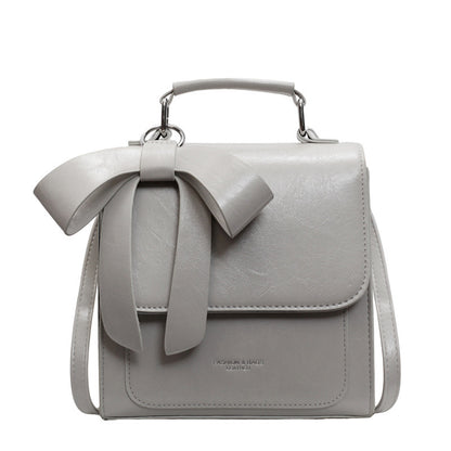 A creamy-white backpack with a bow design on the front, visible straps, and a zipper closure, held in a person's hand.