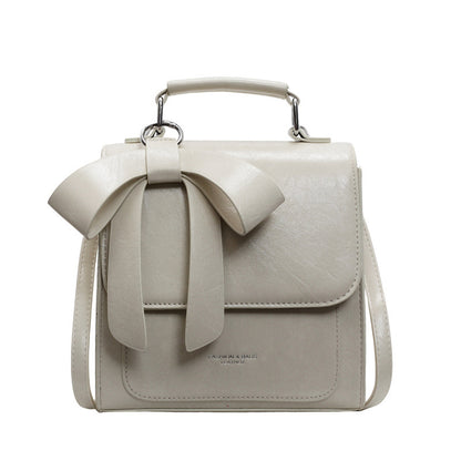 A creamy-white backpack with a bow design on the front, visible straps, and a zipper closure, held in a person's hand.